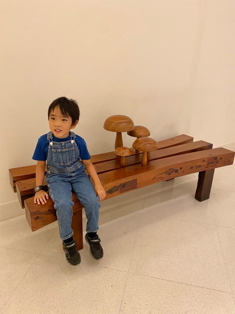 Little boy in denim dungarees sitting on a bench with carved mushrooms
